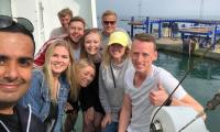 En gruppe unge står sammen og smiler med en havn i baggrunden, hvor vandkanten kan ses.
